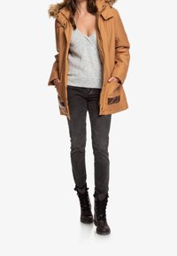 Parka color canela con capucha forrada de piel, llevada sobre un suéter gris de punto trenzado, combinada con jeans gris oscuro y botas negras de cordones.
