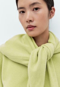 Maglione lavorato a maglia di un verde chiaro, con un fiocco al collo, dalla texture morbida e dal design arrotondato. Primo piano che mostra i dettagli del tessuto.