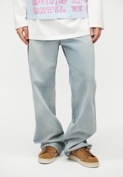Personne portant un jean large bleu clair, une chemise à manches longues blanche, un tablier rayé avec du texte rouge, et des baskets en daim marron avec des semelles blanches.