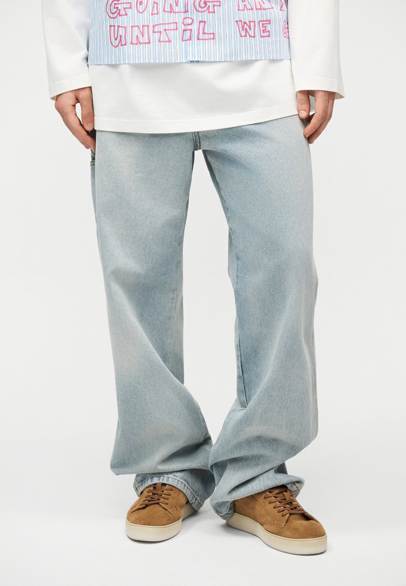 Personne portant un jean large bleu clair, une chemise à manches longues blanche, un tablier rayé avec du texte rouge, et des baskets en daim marron avec des semelles blanches.