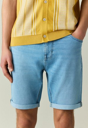 Person trägt hellblaue, umgeschlagene Jeansshorts und ein gelb gestreiftes Hemd mit Knöpfen, eine Hand in der Tasche.