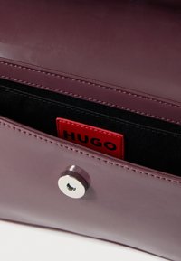Fialová lesklá kabelka so strieborným zapínaním na zacvaknutie. Vo vnútri je čierna textilná podšívka a červená štítok s logom označeným "HUGO".