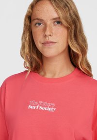 Korallivärviline puuvillane T-särk ümmarguse kaelusega, millel on valged sõnad "Tuleviku Surf Selts" julge fondiga, kerge lõdva istuvusega.