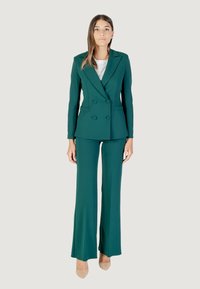 Una mujer se presenta con confianza en un traje verde a medida y tacones beige, con el cabello largo y una blusa blanca, frente a un fondo neutro.