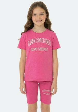 Fille souriante aux longs cheveux ondulés portant un ensemble assorti T-shirt et short Lee Cooper rose vif avec des textes blancs.