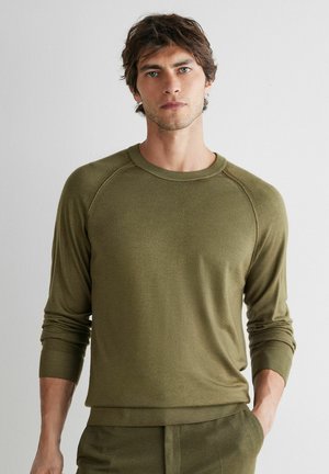 Młody mężczyzna z rozczochraną fryzurą, ubrany w oliwkowozielony sweter z długim rękawem i pasujące spodnie, stojący na jednolitym jasnym tle.