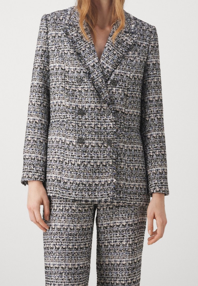 Femme portant un blazer texturé noir, blanc et gris à double boutonnage avec un pantalon taille haute assorti.