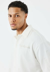 Camisa blanca de botones con un tejido texturizado, que presenta diseños de cactus bordados y un cuello contrastante.