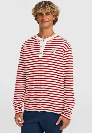 Červenobiele pásikavé tričko s dlhým rukávom, gombíkmi na výstrihu a logovou nášivkou na ľavej strane hrudníka. Mäkká bavlnená látka. Neformálny strih.