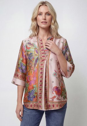 Blond vrouw die een losse, kleurrijke floral zijden blouse met driekwart mouwen en blauwe spijkerbroek draagt, staand tegen een effen witte achtergrond.