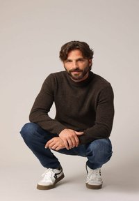 Bruine gebreide trui, blauwe denim jeans en witte sneakers met bruine accenten. Het model hurkt en toont de pasvorm en details van de outfit.