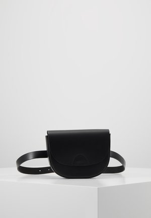 Kleine schwarze Leder-Gürteltasche mit abgerundetem Klappe und verstellbarem Riemen, präsentiert auf einer weißen Eckfläche vor einem schlichten grauen Hintergrund.
