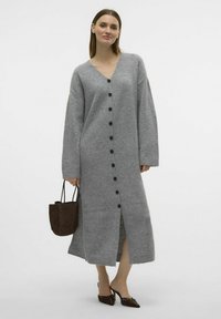 Largo vestido de cárdigan gris hecho de un material suave, con escote en V, mangas largas, botones en el frente y aberturas laterales. Acompañado de un bolso marrón.