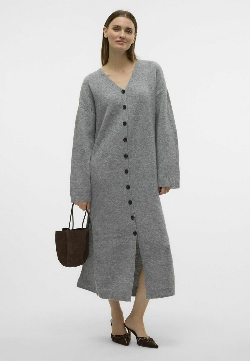 Largo vestido de cárdigan gris hecho de un material suave, con escote en V, mangas largas, botones en el frente y aberturas laterales. Acompañado de un bolso marrón.