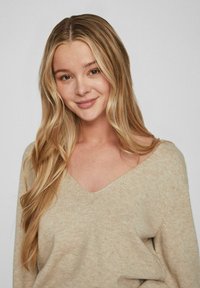 Helles beige V-Ausschnitt-Kapuzenpullover mit langen Ärmeln, gefertigt aus einem weichen, strukturierten Strick, der subtile tonale Variationen im Stoff aufweist.