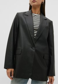 Vrouw in een zwarte nep leren blazer over een zwart-wit gestreept shirt en lichtblauwe jeans, staand tegen een effen achtergrond.