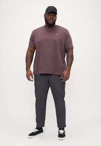 Bordeauxfarbenes T-Shirt mit kurzen Ärmeln und kleinem gelben Logo, kombiniert mit dunkelgrauen Cargo-Hosen und schwarzen Sneakern. Schlichtes Hintergrund.