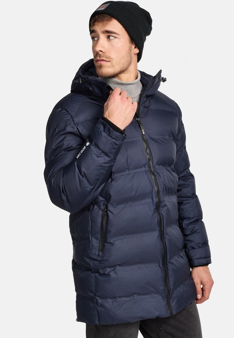 Abrigo acolchado de color azul marino con capucha, que presenta un cierre de cremallera, bolsillos laterales y puños elásticos, fabricado con material ligero y con un diseño acolchado.