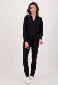 Vrouw met golvend bruin haar draagt een zwarte blouse met lange mouwen en V-hals, bijpassende zwarte broek en witte sneakers, staand tegen een effen achtergrond.