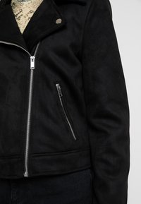 Černá bunda z umělé semiše s krátkým střihem, stříbrným zipem, boční kapsou na zip a klopovým límcem. Hladká textura a minimalistický design.