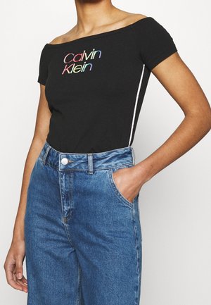 Kobieta w czarnym topie Calvin Klein odsłaniającym ramiona z kolorowym logo i wysokim stanem niebieskich jeansów, jedna ręka w kieszeni.