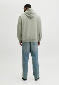 Jasnozielona bluza z dużym kapturem, noszona z luźnymi jasnoniebieskimi dżinsami, widoczna z tyłu, pokazująca swobodny krój i teksturę materiału.