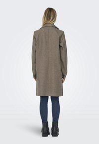 Manteau en mélange de laine marron avec un grand col et des manches longues, doté d’une fente sur les côtés, porté avec un jean skinny bleu foncé et des bottines noires.