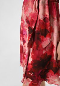 Blumenkleid in Rottönen mit fließendem Stoff. Es zeigt überlappende Blütenblätter in Rosa- und Dunkelrot-Tönen, akzentuiert durch einen seitlichen Knoten.