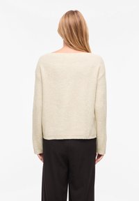 Ελαφρώς μπεζ πλεκτό πουλόβερ με φαρδύ λαιμόκοψη, μακριά μανίκια και κοντό τελείωμα. Απαλή υφή με ριγωτό σχέδιο, φορεμένο πάνω από μαύρο παντελόνι.