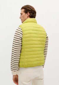 Limoengroene puffer vest met een hoge kraag en een horizontaal gestikt ontwerp. Gstyled over een gestreept, langarm shirt in beige- en bruintinten.