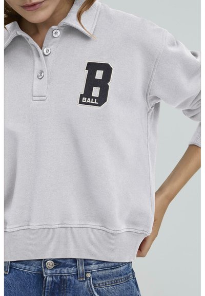 Svijetlo siva dukserica s kragnom, s tri gumba, izvezeni "B BALL" logo u crnoj boji i rebrasti rub. U kombinaciji s plavim trapericama.