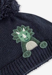 Căciulă tricotată de culoare bleumarin, cu un finisaj texturat, având broderie cu un triceratops verde în față și un pompon pufos în vârf.