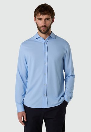 Uomo in piedi che indossa una camicia azzurro chiaro a maniche lunghe con bottoni e pantaloni neri, davanti a uno sfondo semplice.