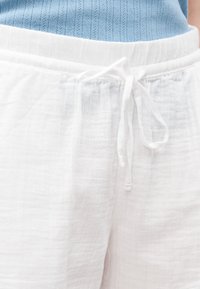 Λευκό υφασμάτινο παντελόνι με ελαστική μέση και δεμένο κορδόνι, φορεμένο με μπλε πλεκτό τοπ.
