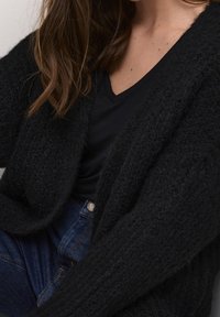 Musta, teksturoidulla pinnalla varustettu neuletakki, jossa on avoin etuosa, ylikokoinen istuvuus ja lasketut hartiat, yhdistettynä mustaan T-paitaan ja farkkuihin.