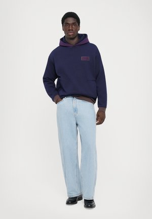 Navy hoodie with a purple interior, featuring a front pocket and logo patch, paired with light blue wide-leg jeans and black shoes.