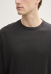 Joven vestido con un suéter de cuello redondo gris oscuro, mostrado desde los hombros hasta la barbilla, mirando ligeramente hacia un lado sobre un fondo claro.