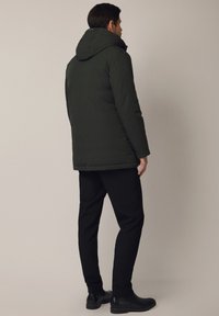 Mężczyzna stojący tyłem, ubrany w ciemnozieloną kurtkę z kapturem, czarne spodnie i czarne skórzane buty, na jednolitym beżowym tle.