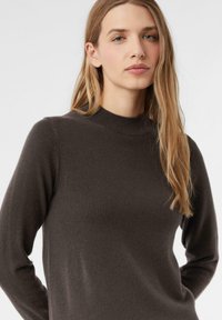 Brauner Strickpullover mit hohem Kragen, langen Ärmeln und taillierter Silhouette. Weiche Textur mit minimalen Details und glatter Oberfläche.