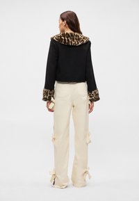 Casaco preto curto com colarinho e punhos de pele sintética com padrão de leopardo, combinado com calças beijas com laços decorativos na bainha.