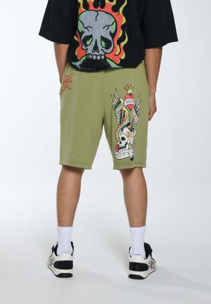 Olive green shorts made of cotton, featuring a colorful tattoo-style graphic of a skull and snake on the side. Worn with a black shirt.
