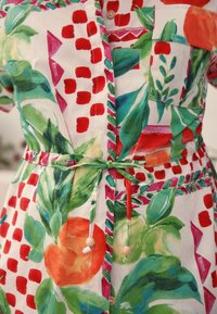 Virágmintás ruha élénk színekkel, nagy zöld levelekkel, piros pöttyökkel és narancssárga díszítésekkel. A deréknál fa gyöngyökkel köthető meg.