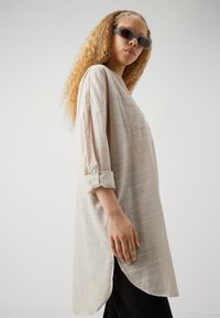 Chemise longue rayée beige clair avec une poche poitrine, des manches retroussées et un ourlet arrondi. Portée avec un pantalon sombre et de grandes lunettes de soleil.