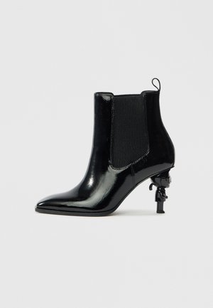 Black patent leather ankle boot with elastic side panels, pointed toe, and a unique sculptural heel design featuring a small figure.