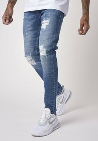 Light blue ripped denim jeans feature varying distress patterns and a slim fit. Paired with white and gray athletic shoes.