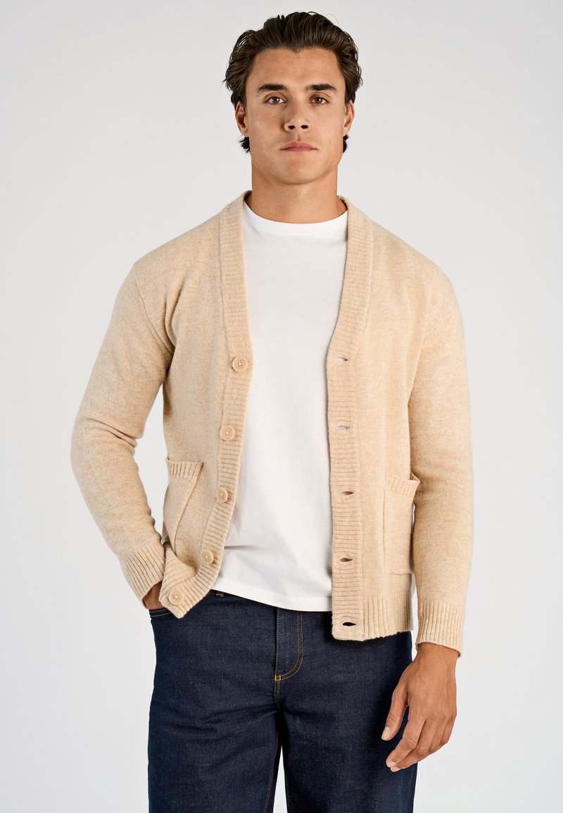 Beige stickad kofta med sjalkrage, fem knappar, ribbade ärmslut och två framfickor, bärs över en vit t-shirt och mörka jeans.