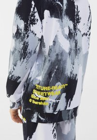 Felpa a maniche lunghe con stampa astratta in nero, bianco e grigio con testo giallo; tessuto morbido, vestibilità comoda, polsini e orlo a coste.