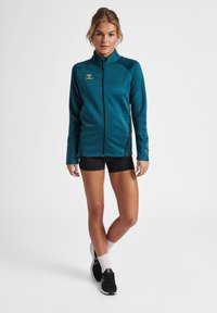 Jasnozielona polarowa kurtka z wysokim kołnierzem, przednim zamkiem i czarnymi akcentami. Posiada logo na klatce piersiowej oraz boczne kieszenie. Noszona z czarnymi szortami.