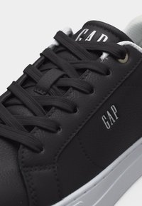 Baskets en cuir noir avec des lacets noirs, logo GAP blanc sur le côté et la languette, et une semelle en caoutchouc blanche.