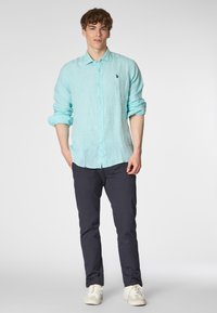 Camicia di lino azzurra chiara con maniche lunghe, chiusura con bottoni e un piccolo logo. Abbinata a pantaloni scuri e scarpe da ginnastica bianche.